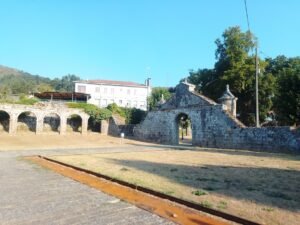 Fachada de piedra y portalón de entrada de las ruinas del Monasterio de Santa María de Melón en Ourense, mostrando las arquerías laterales y un gran patio de tierra en un día despejado.