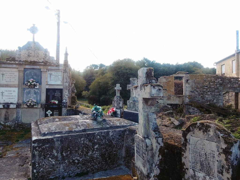 • Cruces de piedra antiguas cubiertas de musgo y panteones familiares en el cementerio parroquial de Santiago de Corneda, rodeado de un espeso bosque de fondo.