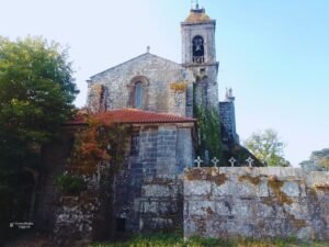 Vista lateral exterior de la iglesia y la torre campanario de piedra del Monasterio de Santa María de Melón en Ourense, Galicia, mostrando un muro antiguo con pequeñas cruces grabadas en un día despejado.