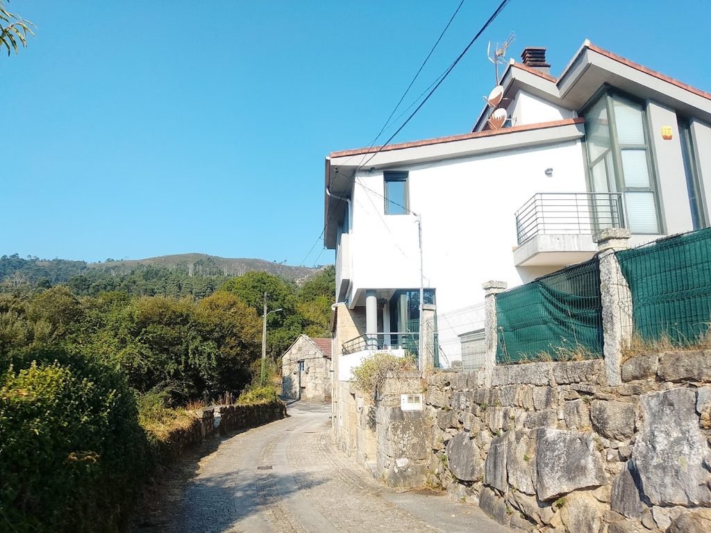 Fachada de una casa residencial de estilo moderno en el pueblo de Melón, Galicia, mostrando dos antenas parabólicas satelitales instaladas bajo un cielo despejado.